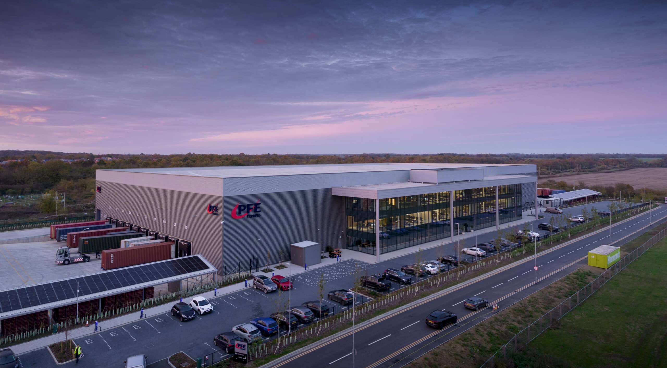 A large warehouse and distribution center with trucks at loading bays, a parking lot with cars, and a road running alongside the building, photographed at dusk.