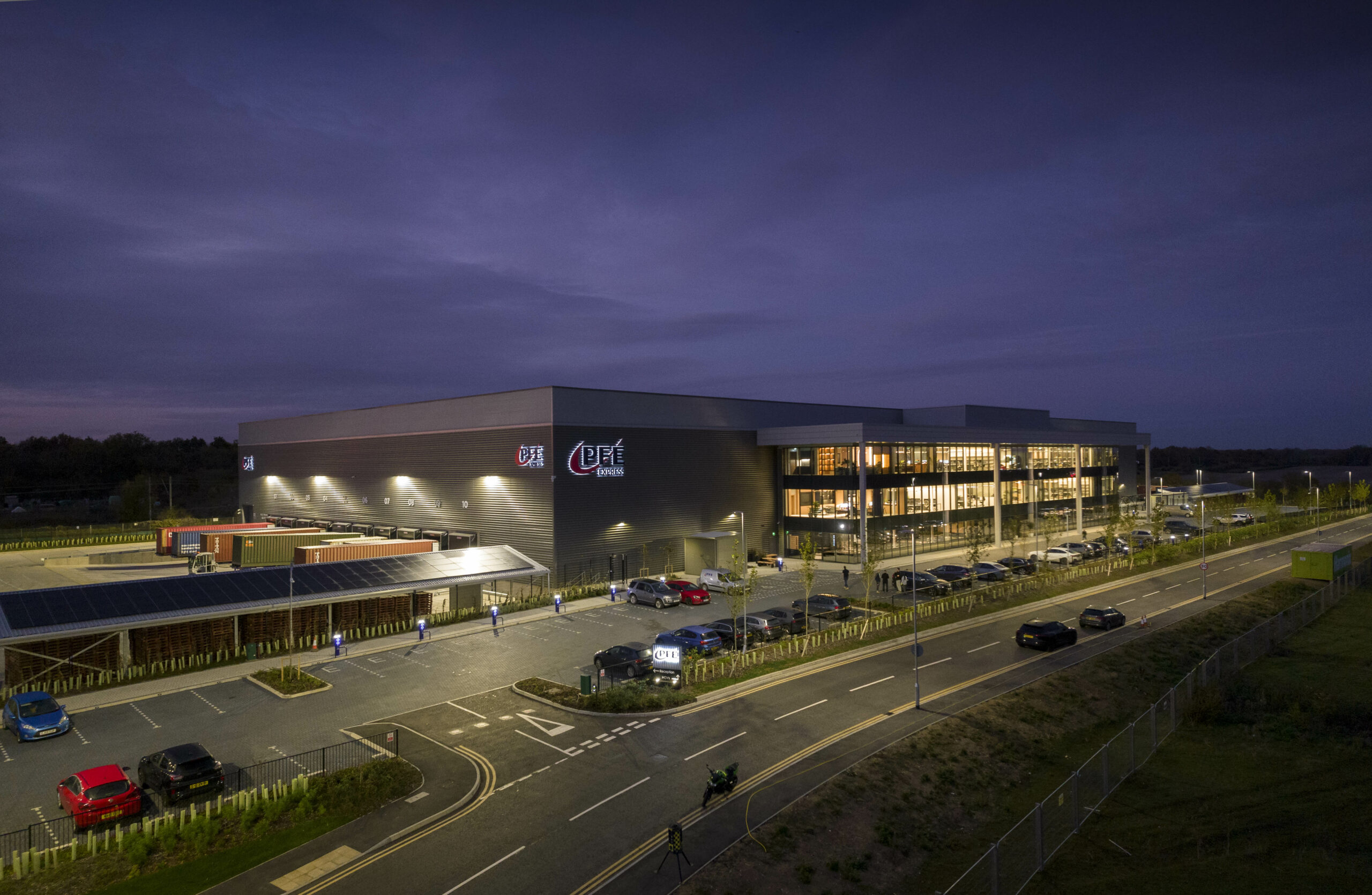 An illuminated modern commercial building with large windows and a parking lot at dusk, cars parked outside, and streetlights along an adjacent road.