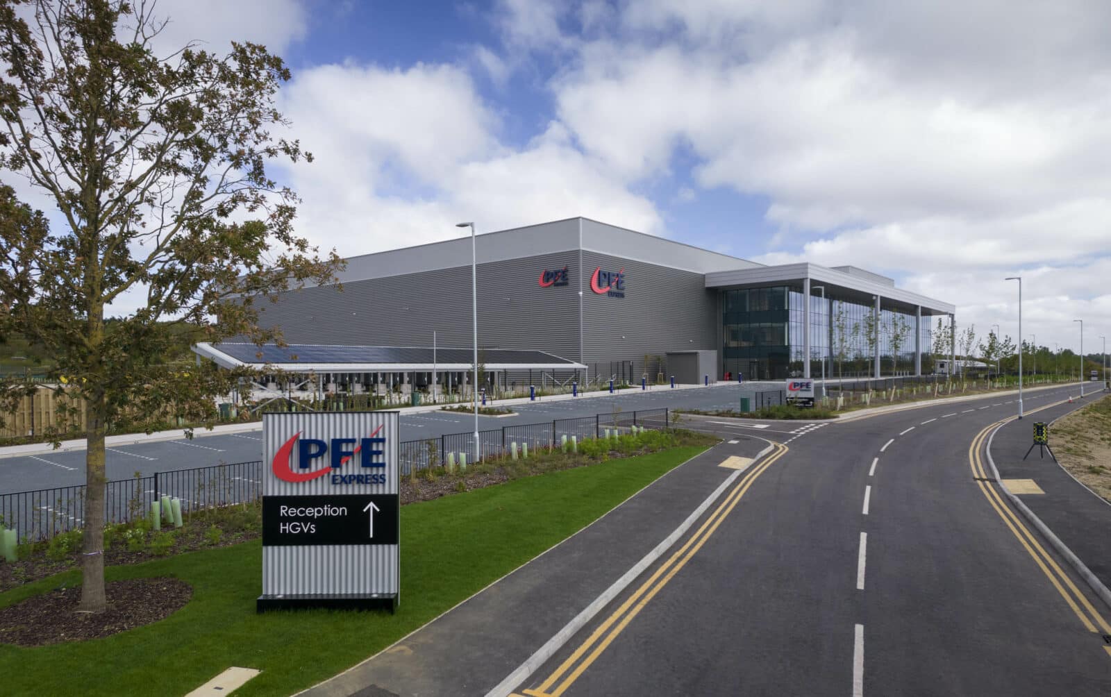 Large modern industrial building with "PFE" signage, a parking area, and a road leading to the entrance; a sign indicates directions for reception and HGVs.