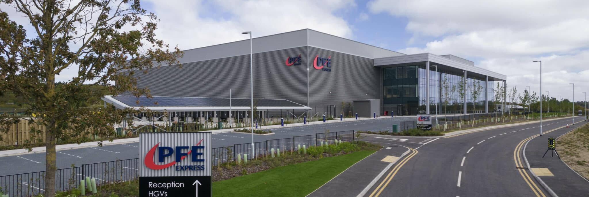 Large modern industrial building with "PFE" signage, a parking area, and a road leading to the entrance; a sign indicates directions for reception and HGVs.