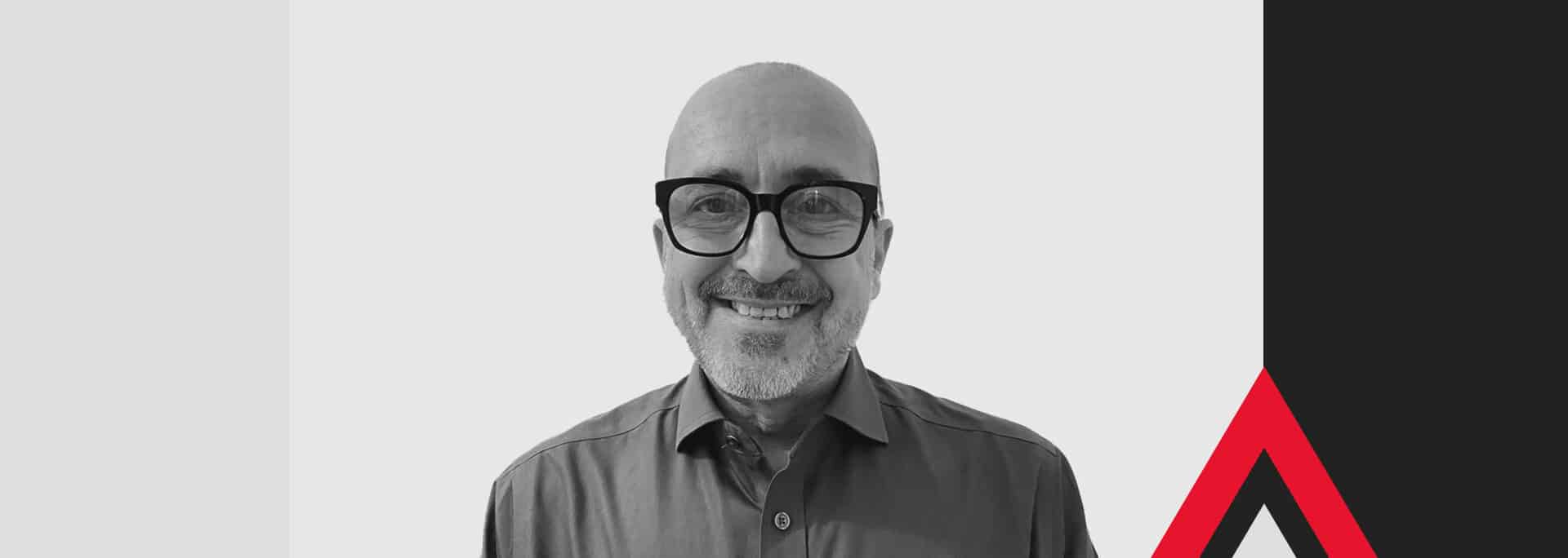 A man with glasses and a collared shirt smiles at the camera, set against a geometric background with maroon, black, and white shapes and an angular red accent.