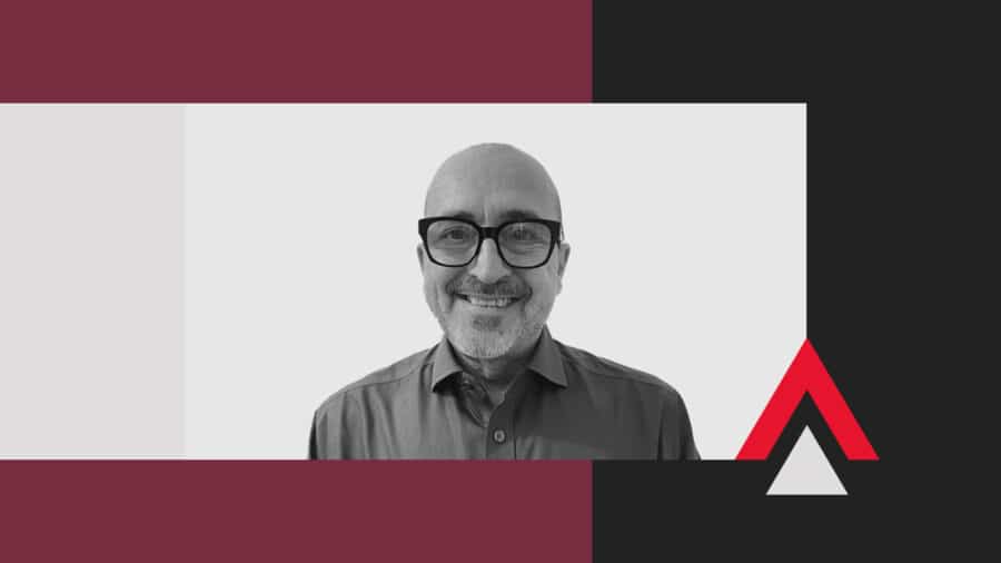 A man with glasses and a collared shirt smiles at the camera, set against a geometric background with maroon, black, and white shapes and an angular red accent.