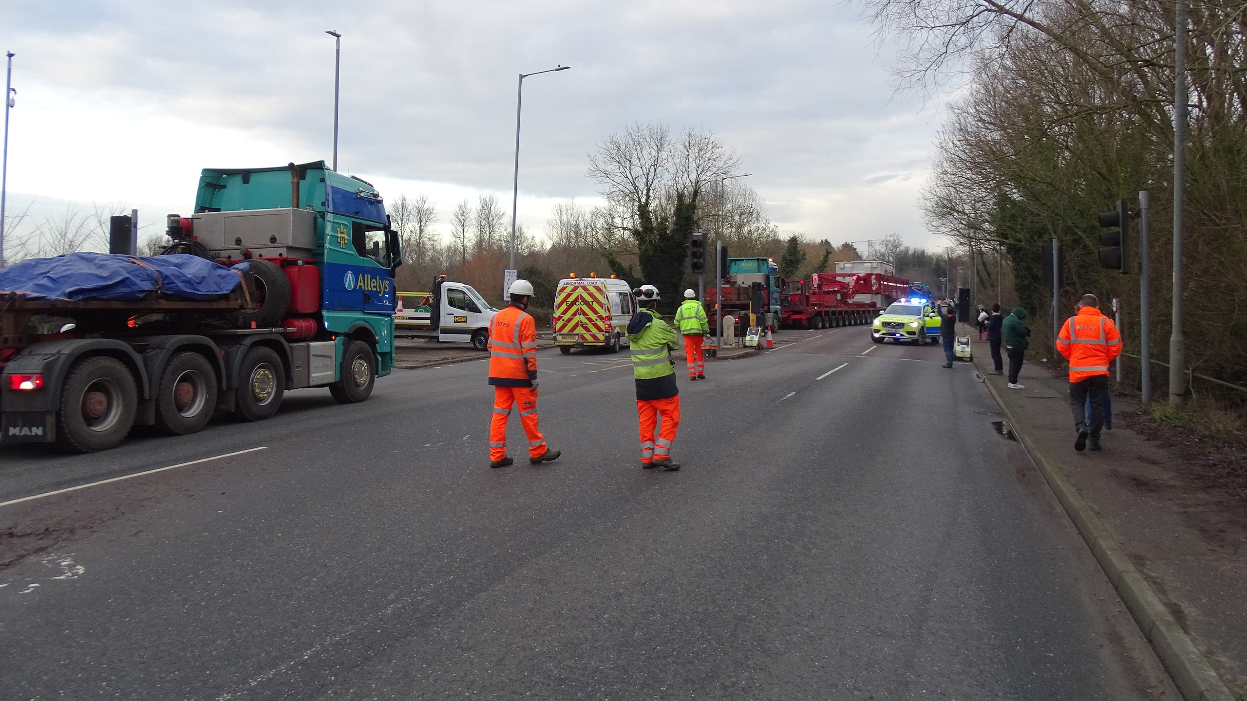 Several workers in high-visibility gear manage traffic around stopped trucks and a police car on a multi-lane road, with some people standing on the roadside.