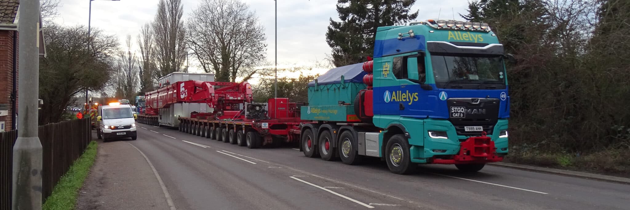 A large Allelys-branded truck transports oversized industrial equipment on a multi-axle trailer along a suburban road, with an escort vehicle ahead.