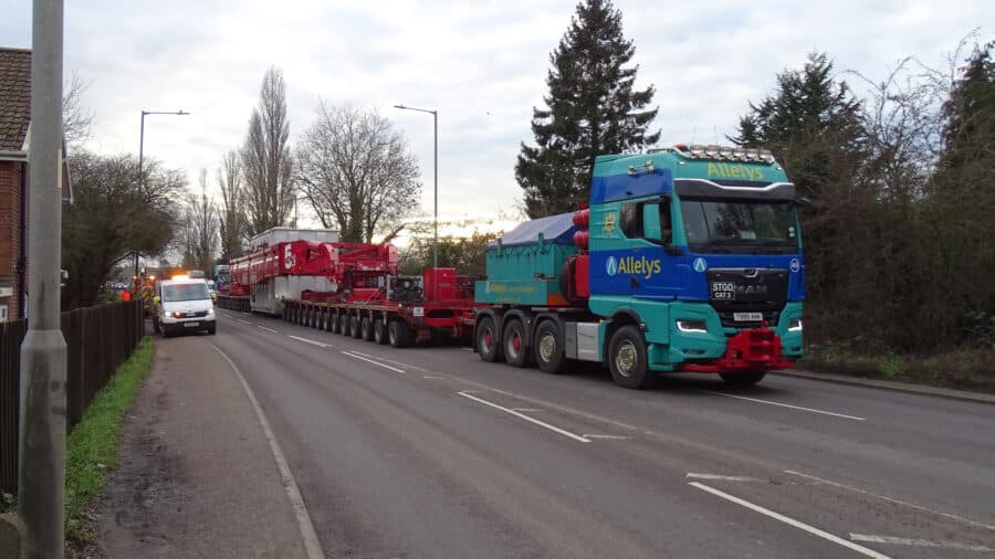 A large Allelys-branded truck transports oversized industrial equipment on a multi-axle trailer along a suburban road, with an escort vehicle ahead.