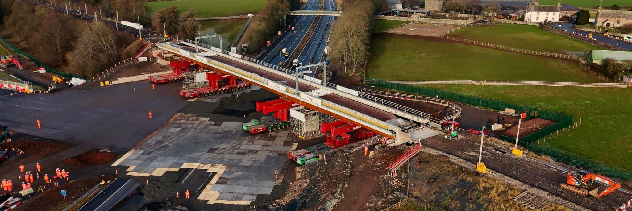 Aerial view of a bridge under construction over the West Coast Main Line and a multi-lane road in a rural area, with workers, machinery, and surrounding green fields visible.