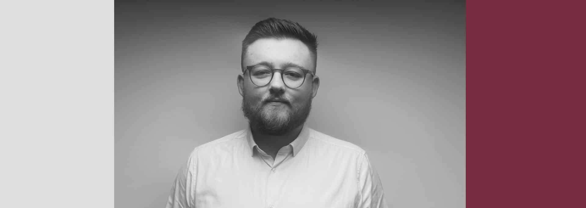 A man with glasses and a beard poses in front of a plain background, framed by geometric color blocks in black, white, and burgundy.