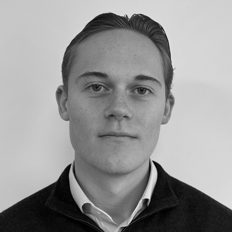 A young man with short, dark hair is facing the camera with a neutral expression, wearing a collared shirt and sweater. The photo is in black and white.