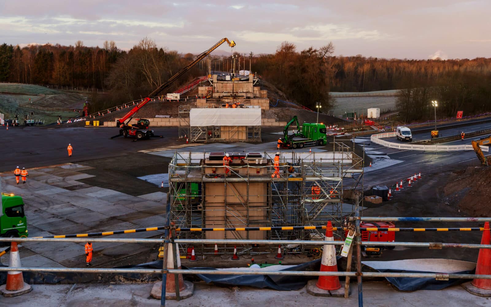 Construction workers and machinery are building a bridge over the West Coast Main Line, with scaffolding, barriers, and equipment visible at the site during the day.