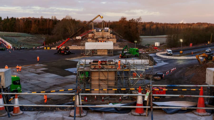 Construction workers and machinery are building a bridge over the West Coast Main Line, with scaffolding, barriers, and equipment visible at the site during the day.