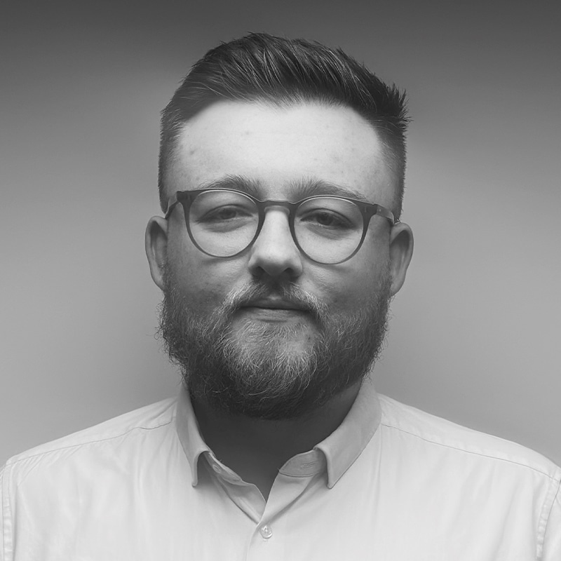 A bearded man with glasses and short hair, wearing a buttoned-up light shirt, faces the camera against a plain, gradient background.