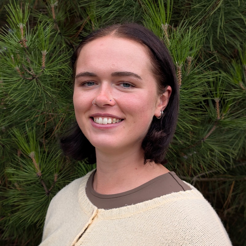 Olivia Blythe, Air Quality Consultant at Create Consulting Engineers, smiling outdoors against evergreen foliage.