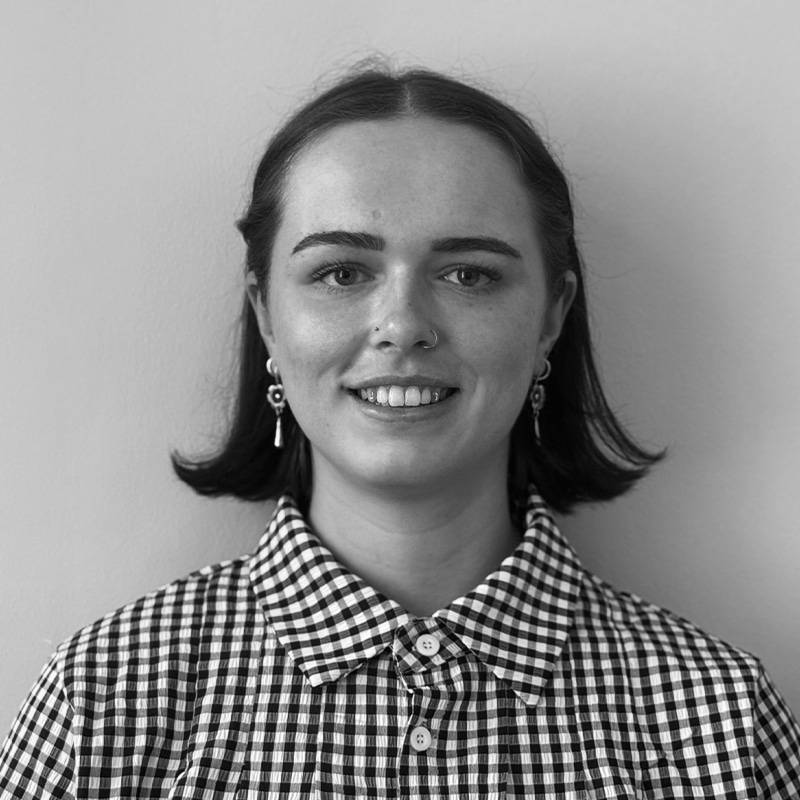 A person with short dark hair, earrings, and a nose ring, wearing a checked shirt, smiles at the camera against a plain background.
