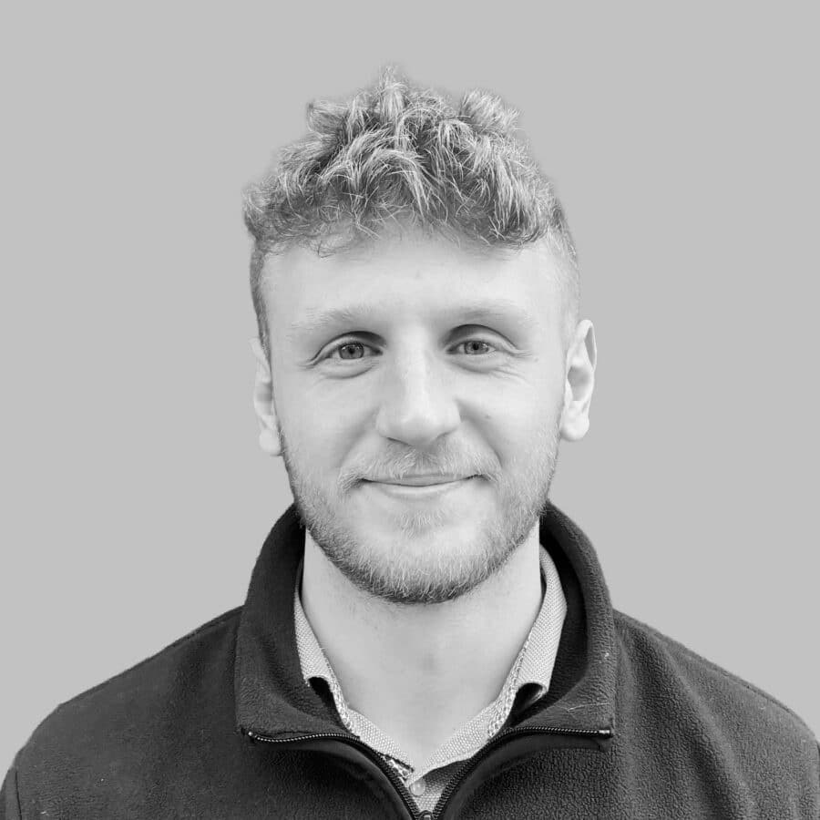 A young man with short curly hair and a beard, wearing a zip-up jacket and collared shirt, faces the camera and smiles slightly. The background is plain grey.