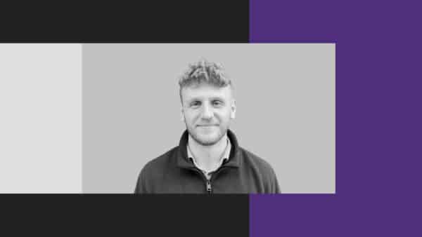 A man with short curly hair and a slight smile faces the camera. The background features a greyscale centre with black, grey, and purple sections on the sides.