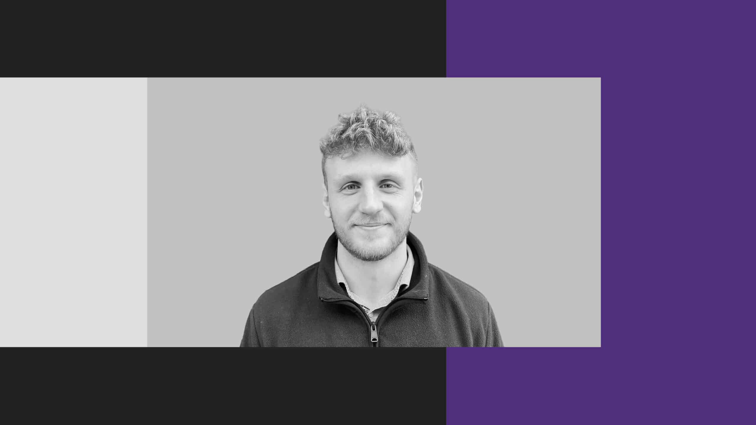 A man with short curly hair and a slight smile faces the camera. The background features a greyscale centre with black, grey, and purple sections on the sides.