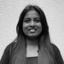 A woman with long dark hair smiles at the camera whilst standing in front of a textured white wall.