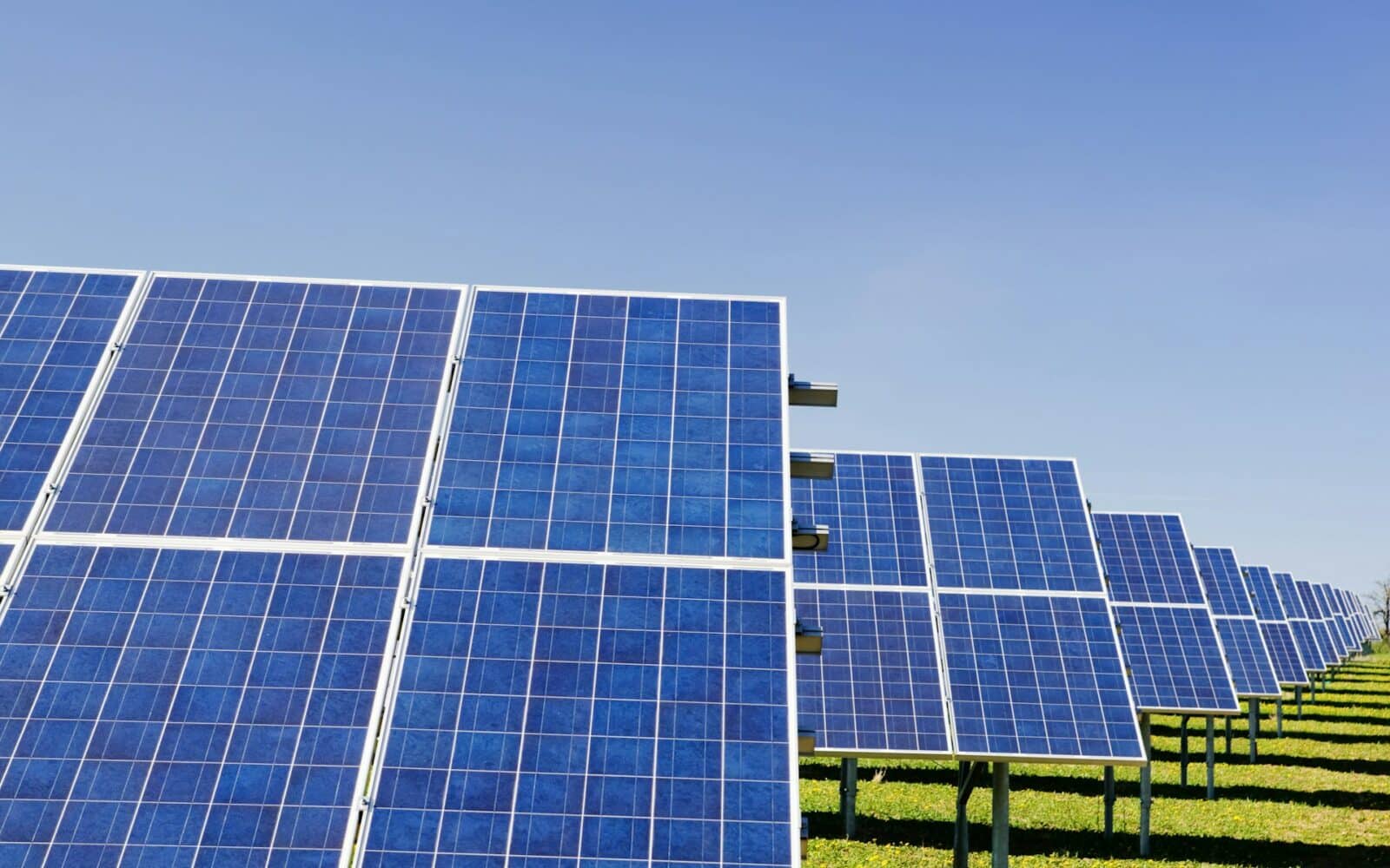 Large-scale photovoltaic solar farm with multiple blue panel arrays mounted on metal frames across green fields under clear sky.