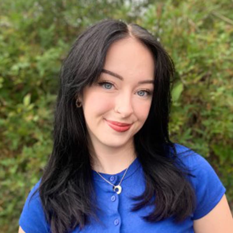 Lola Rogerson, team member at Create Consulting Engineers, wearing blue shirt against green foliage background.