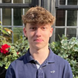 Portrait of a young male team member at Create Consulting Engineers, wearing navy polo shirt, positioned outdoors with greenhouse and garden visible behind.