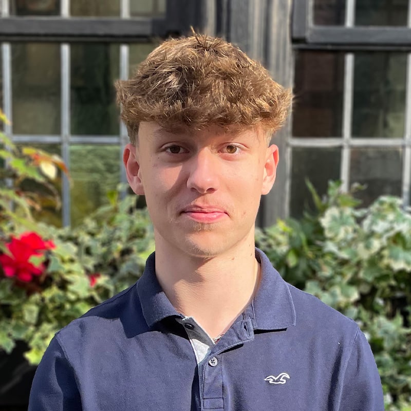 Portrait of a young male team member at Create Consulting Engineers, wearing navy polo shirt, positioned outdoors with greenhouse and garden visible behind.