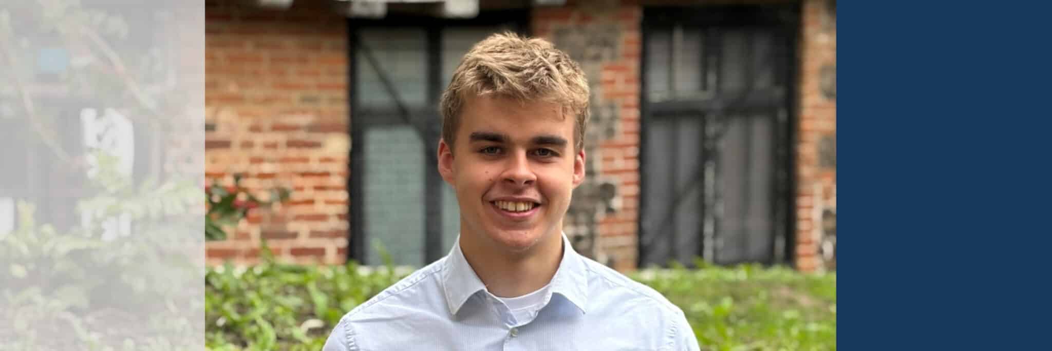 Professional headshot of a young man in light blue shirt, smiling at camera against brick building background.