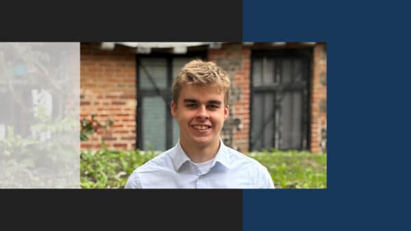 Professional headshot of a young man in light blue shirt, smiling at camera against brick building background.
